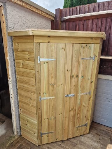 Wooden storage shed with a sloping roof and double doors, placed against a fence.