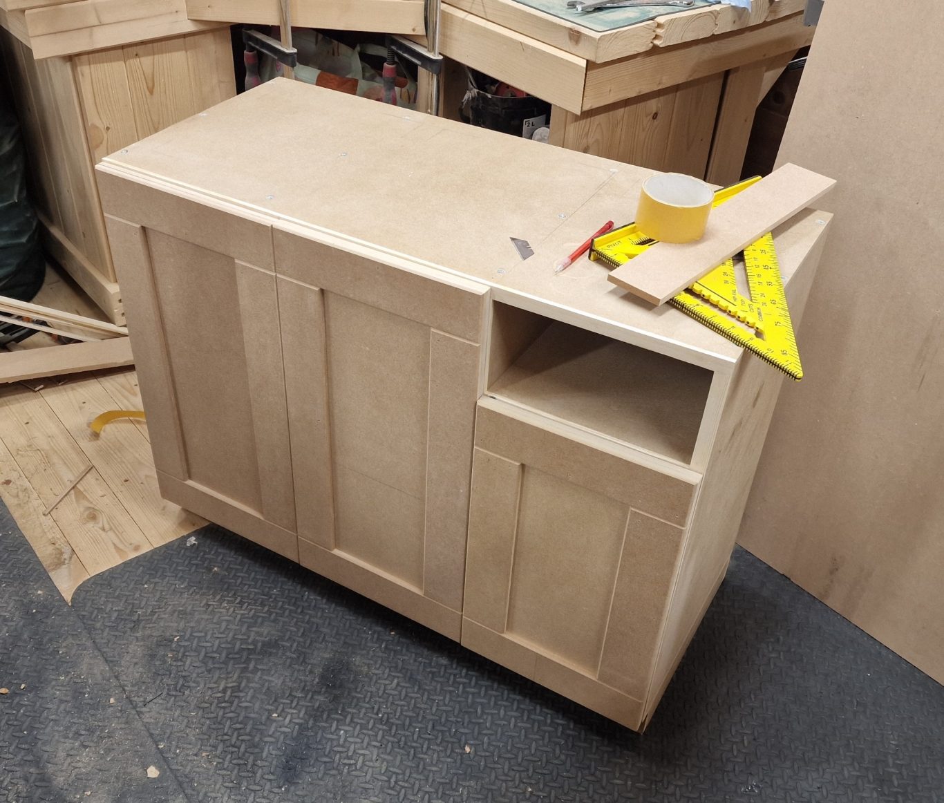 A bare wooden cabinet with an open shelf and adjacent small drawer, amid a workshop.