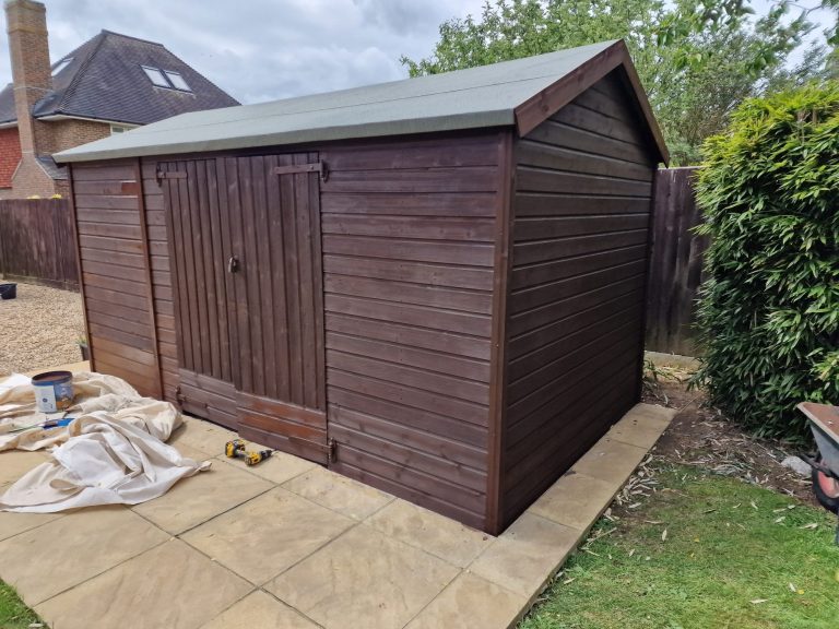 Brown wooden garden shed with a sloped roof, set on a paved patio.